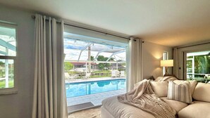 A living room with a large window overlooking a swimming pool and outdoor seating area. - Beautiful Gulf Access Home with Heated Pool and Private Dock in Yacht Club (Cape Coral)