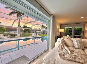 Una habitación con un amplio ventanal con vistas a una piscina y al atardecer.
