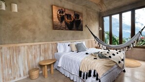 A bedroom with a hammock, a bed with a white and beige quilt, a small wooden stool, and a painting of boats on the wall. - Pousada Zinga Caraíva (Porto Seguro)