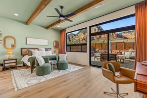 A spacious bedroom with a large bed, two green ottomans, a desk, and a chair. A ceiling fan and a large window with a view of a rocky landscape are visible. - Mystic Mesa | Heated Pool, Spa, Firepit, Views (Sedona)