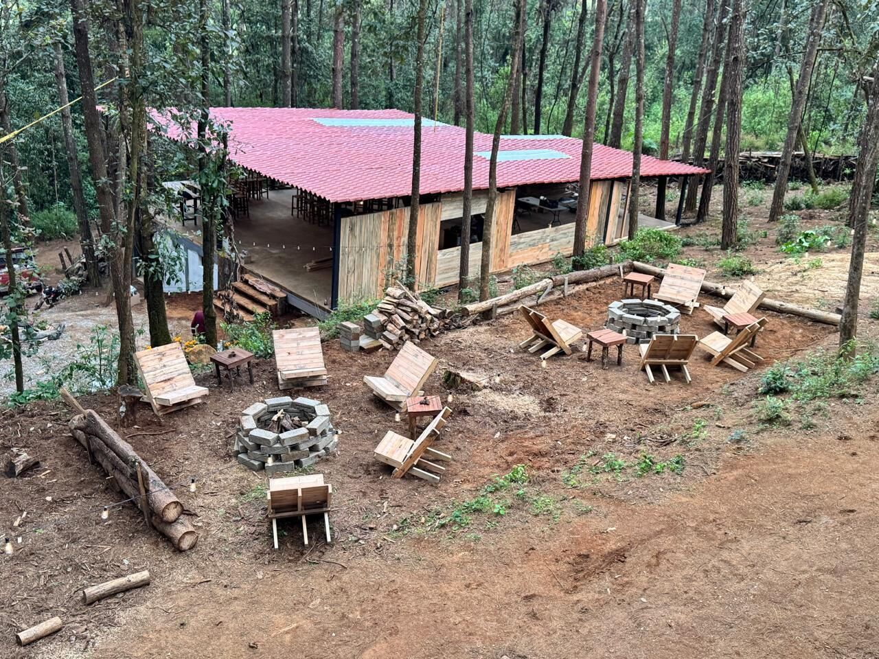 Una cabaña rústica con techo rojo, rodeada de un bosque. Hay varias sillas de madera y un hoyo para la fogata.