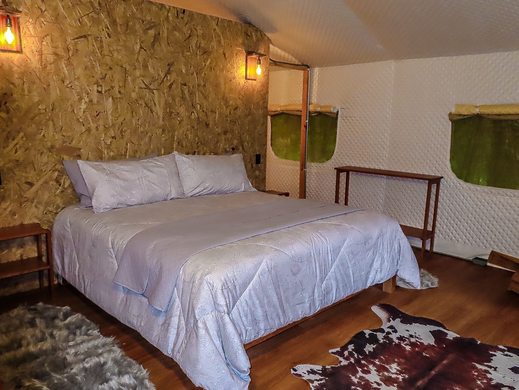Un dormitorio con pared de piedra, cama con colcha estampada, mesita de noche de madera, alfombra de cuero de vaca y dos lámparas fijadas a la pared.