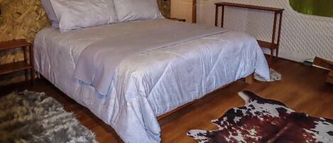 Un dormitorio con pared de piedra, cama con colcha estampada, mesita de noche de madera, alfombra de cuero de vaca y dos lámparas fijadas a la pared.