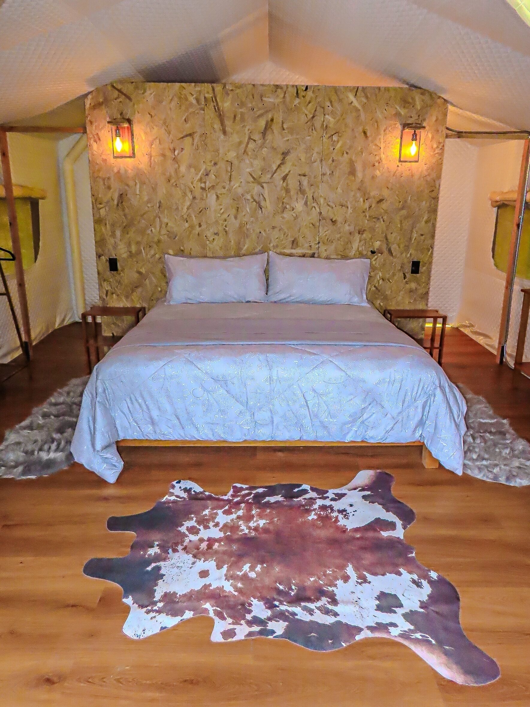 A bedroom with a stone-textured wall, two bedside lamps, a bed with a light blue bedspread, and a large animal hide rug on the floor.