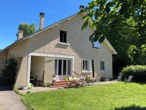 Una casa de dos pisos con un porche cubierto, una chimenea y un pequeño jardín con muebles de exterior.