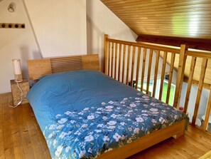 Quarto com estrutura de cama de madeira, colcha florida azul, painel de cabeceira de madeira, mesa de cabeceira de vime com luminária e vista para o exterior através de uma janela.