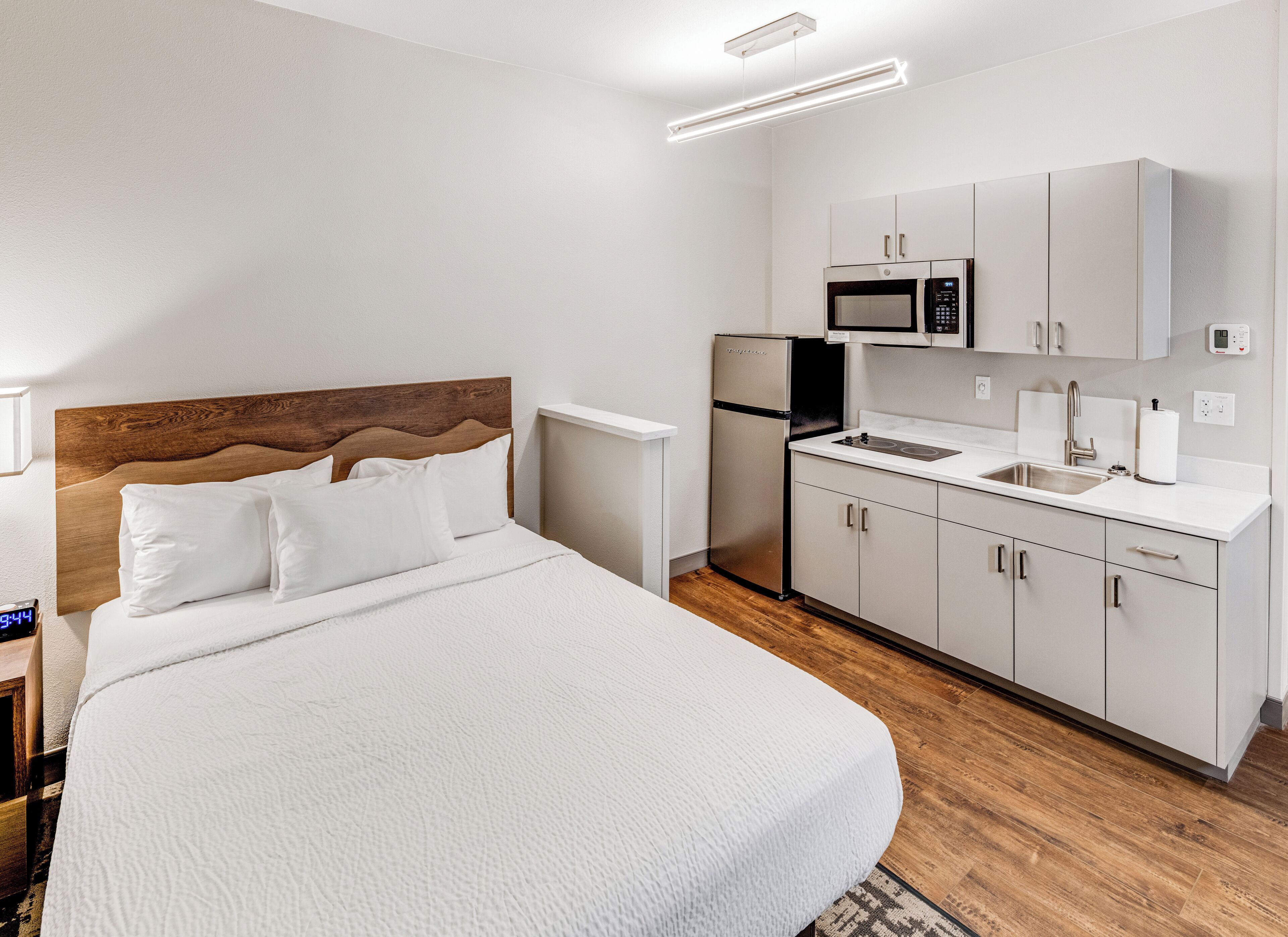Una camera da letto con un letto, un comodino, una sveglia digitale, una cucina con forno a microonde, frigorifero, lavello e pensili.