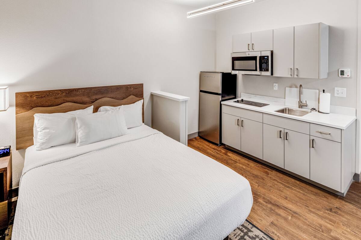 Ein Schlafzimmer mit einem Bett, einem Nachttisch, einer digitalen Uhr, einer Küche mit Mikrowelle, Kühlschrank, Spüle und Schränken.