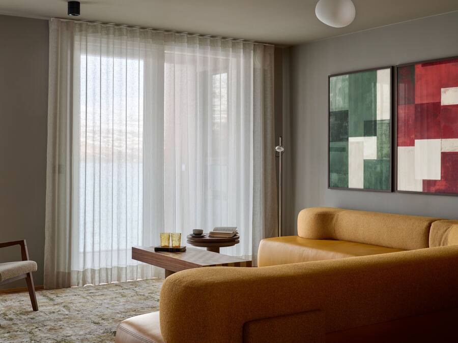 A living room with a sofa, a chair, a table with a book and glasses, and a window with sheer curtains.