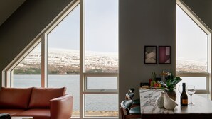 Ein Zimmer mit einem großen Fenster mit Blick auf ein Gewässer, ein braunes Ledersofa, ein Esstisch mit Stühlen und eine kleine Pflanze.