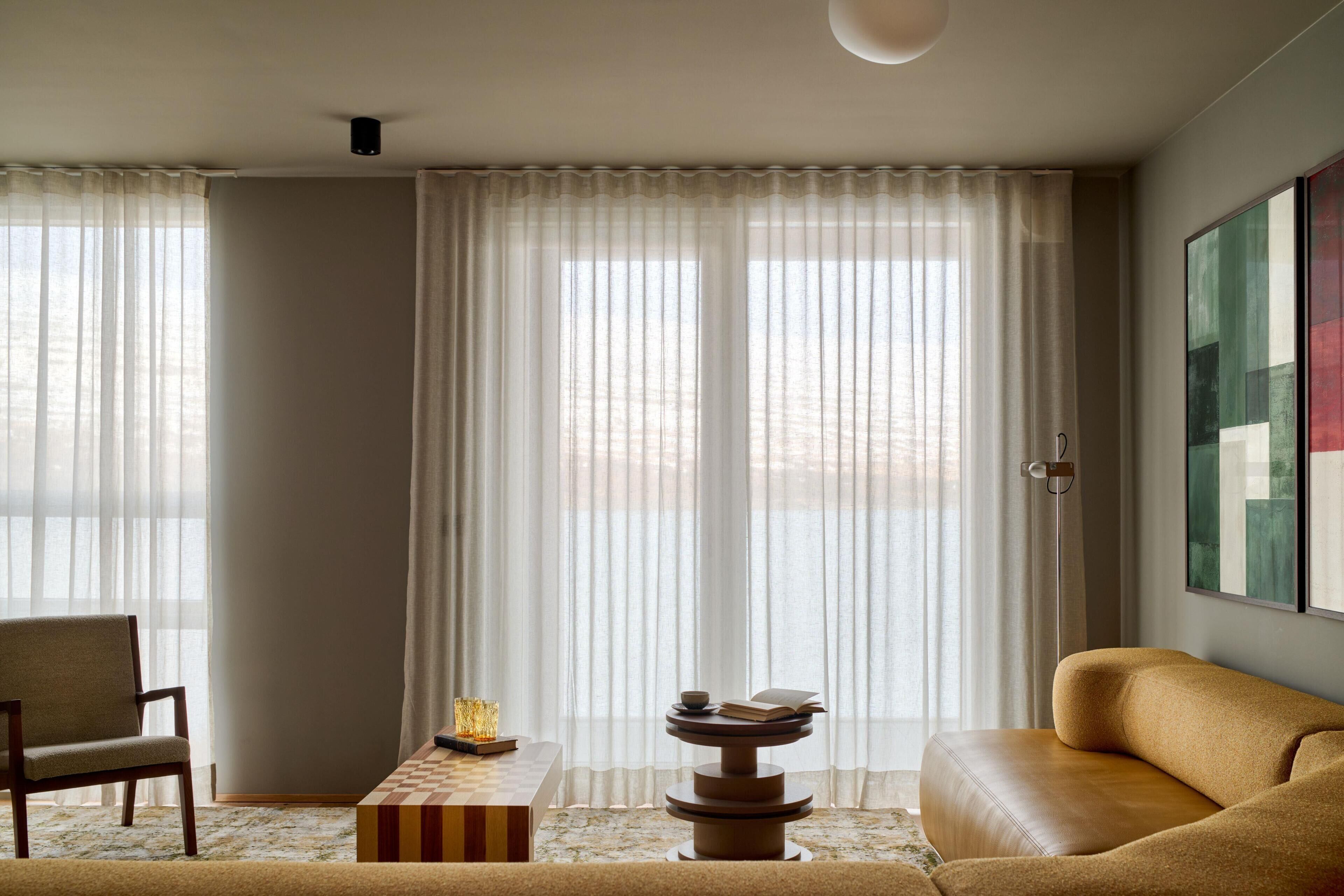 Una sala de estar moderna con un sofá, una mesa de centro de madera y una silla. Hay un ventanal con cortinas translúcidas y un cuadro en la pared.