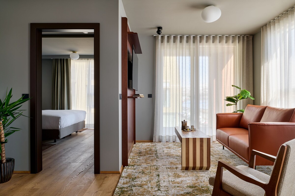 A modern hotel room with a bed, a sofa, a coffee table, and a large window with sheer curtains.