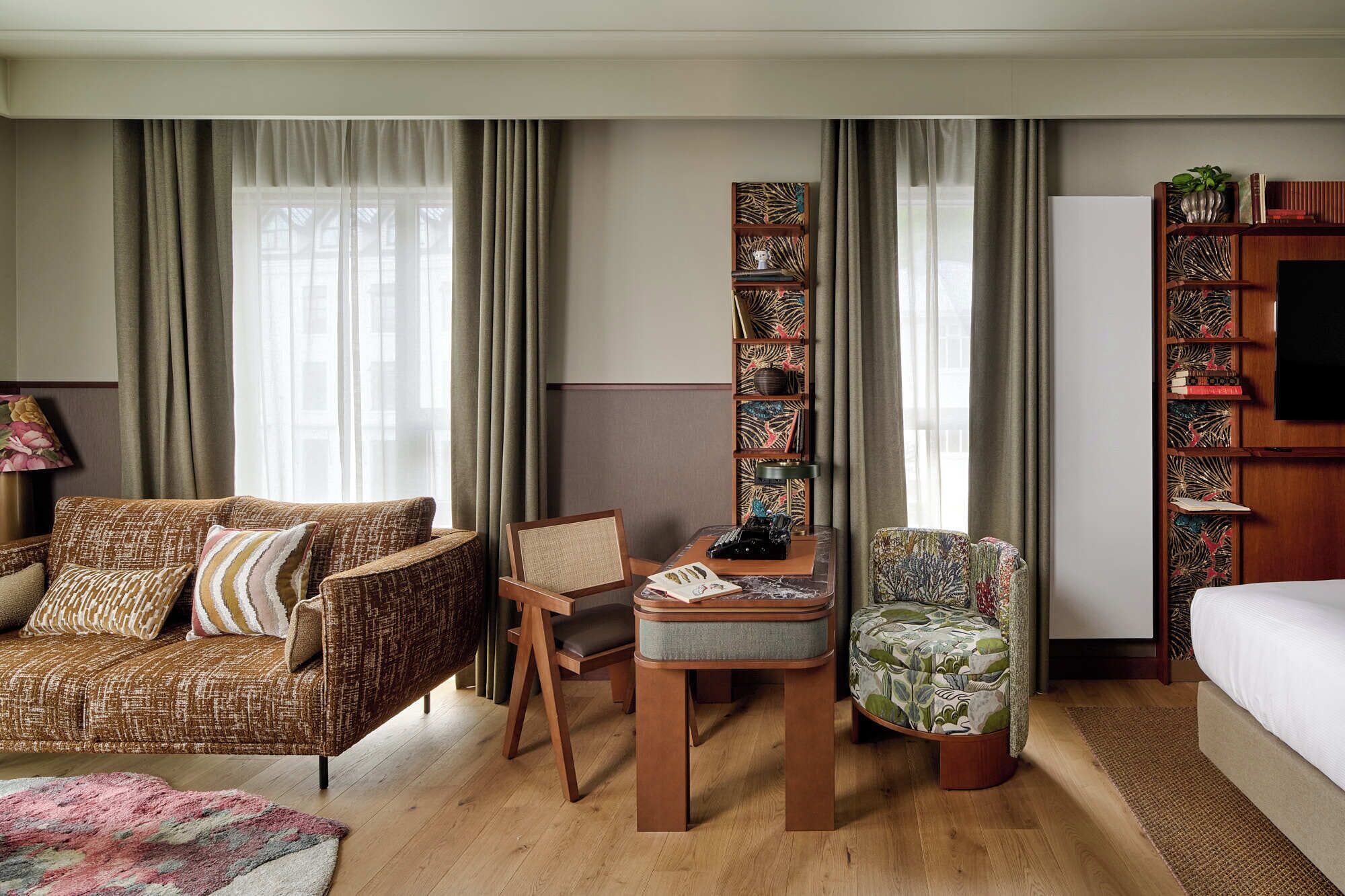 A hotel room with a sofa, armchair, small table, bookshelf, and a bed.