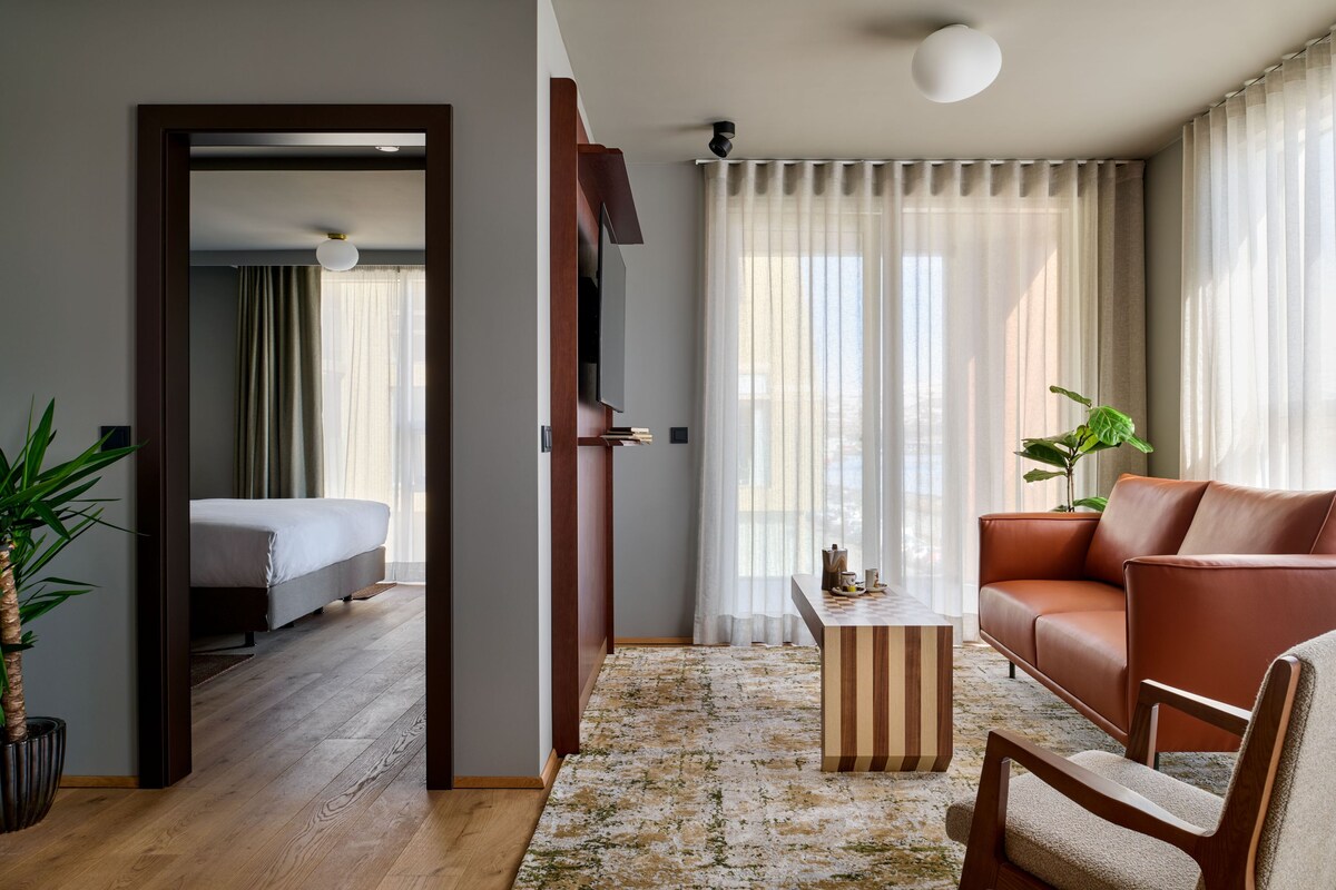 A modern hotel room with a bed, a sofa, a coffee table, and a large window with sheer curtains.