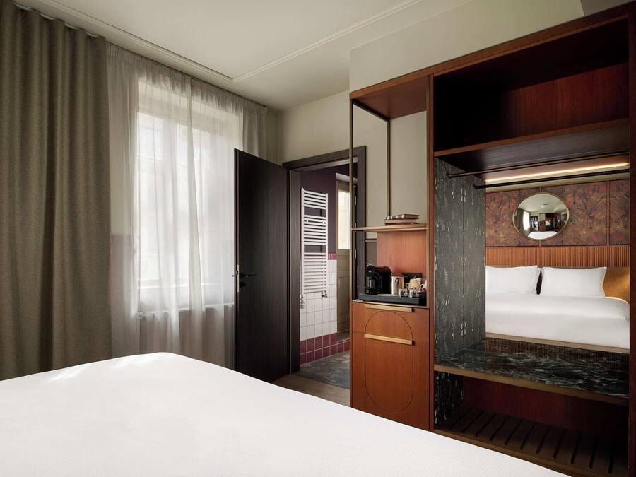 Una habitación de hotel con cama, escritorio, espejo y vista a un pasillo.