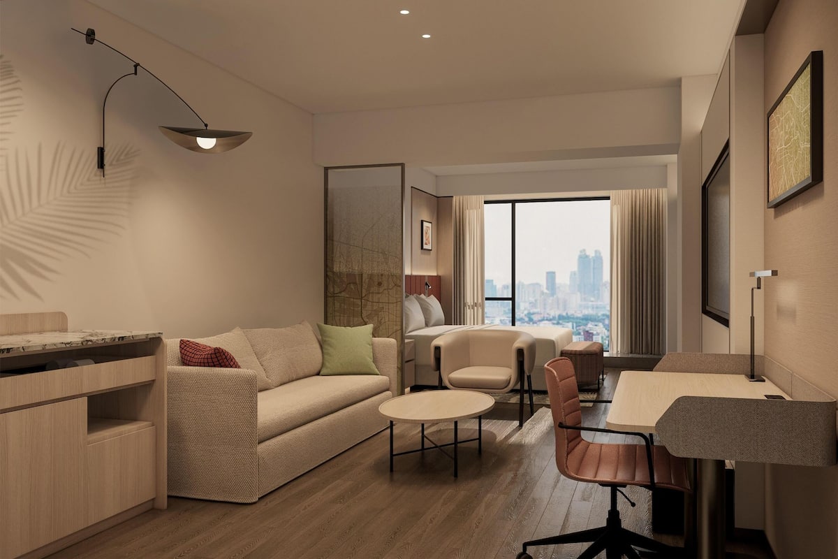 A modern hotel room with a sofa, a dining table, and a city view.
