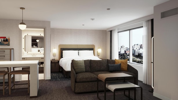 Habitación de hotel moderna con una cama grande, un sofá, un escritorio con dos sillas, vista a la ciudad y un baño con espejo y lavamanos.