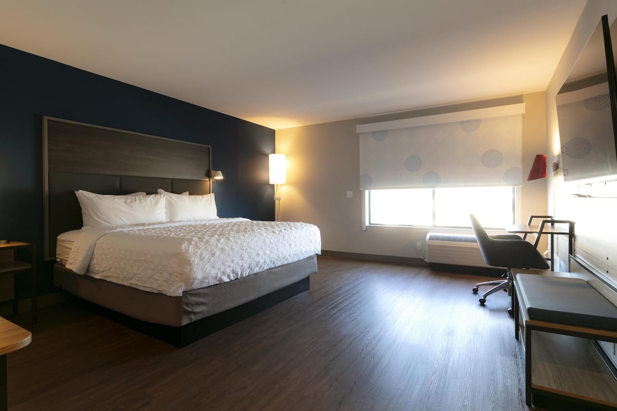A hotel room with a bed, a desk, a chair, a window with blinds, and a wall-mounted TV.