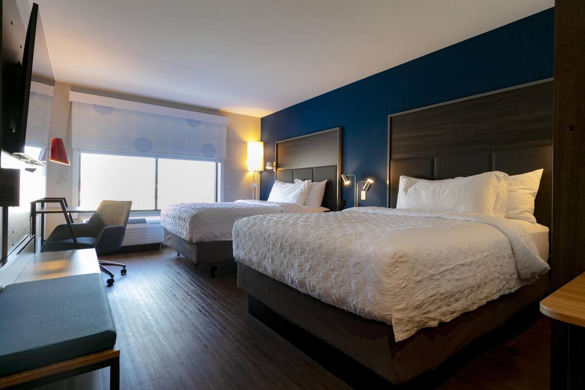 A hotel room with two beds, a desk, a chair, and a TV.