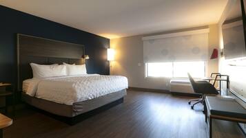 A hotel room with a bed, a desk, a chair, a window with blinds, and a wall-mounted TV.