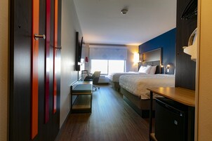 Habitación de hotel con cama, escritorio, silla, refrigerador pequeño y ventana con persianas.