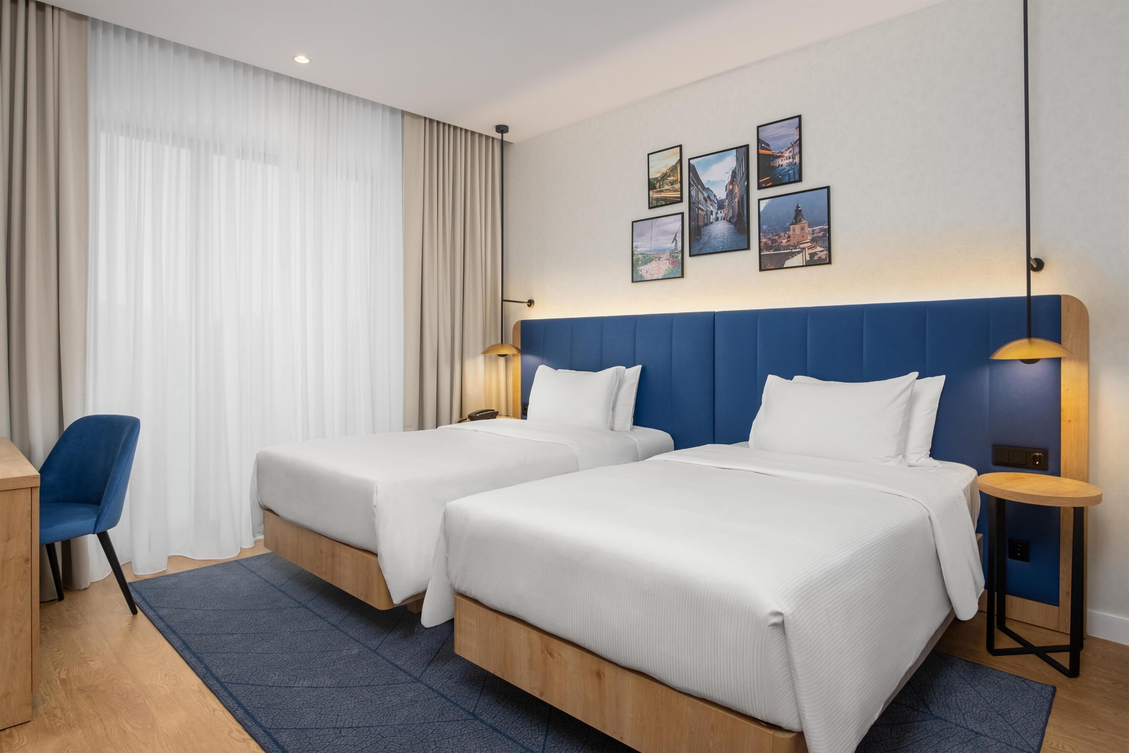 A hotel room with two beds, a desk, a chair, and a large window with sheer curtains.