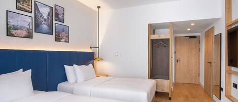 A hotel room with two beds, a blue headboard, a wall with framed pictures, a hanging shower, and a wooden wardrobe.