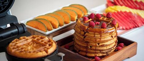 A waffle maker with waffles and a stack of pancakes with raspberries and syrup.