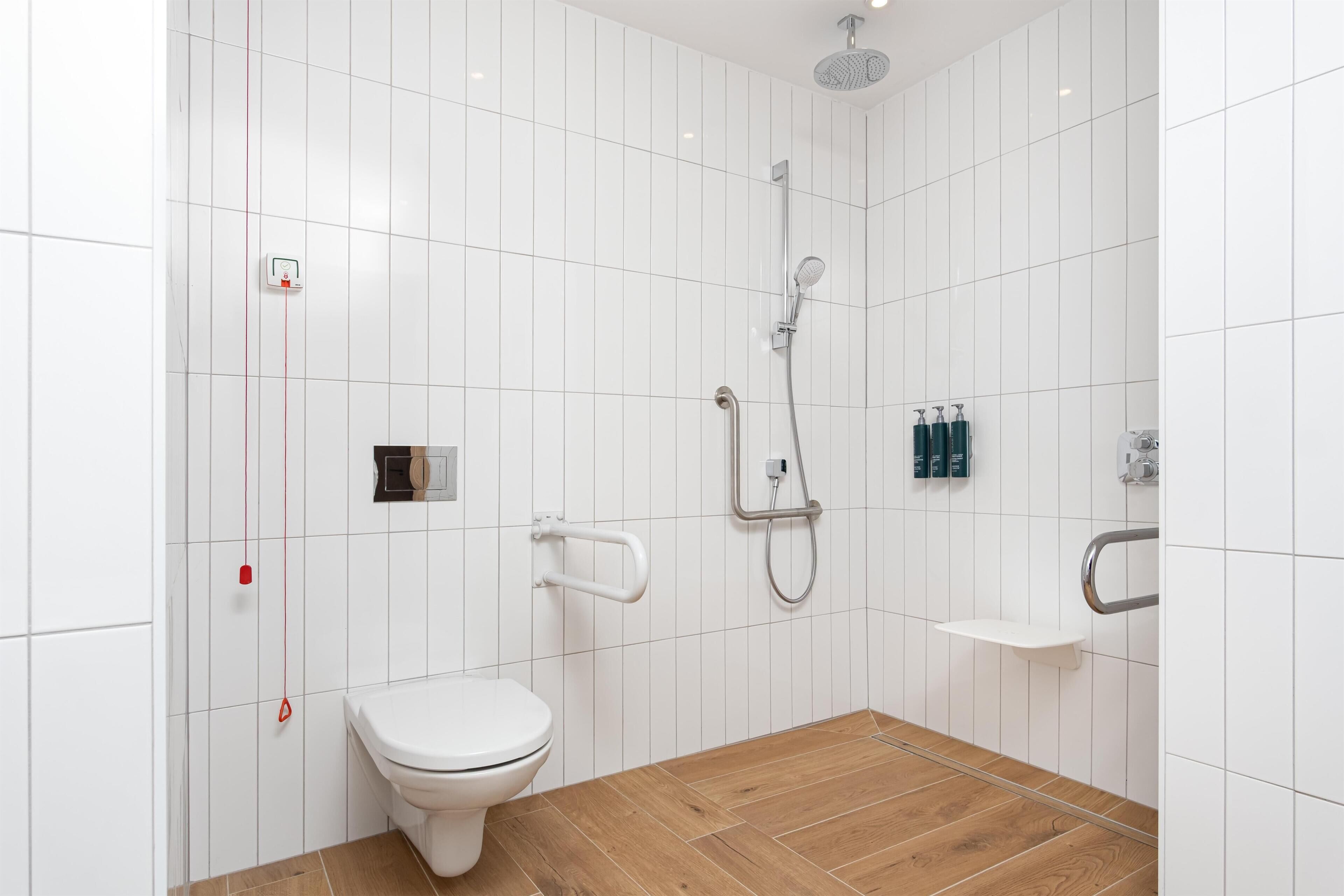 A modern bathroom with white tiled walls, a wooden floor, a toilet, a shower with a handheld showerhead, and a small shelf.