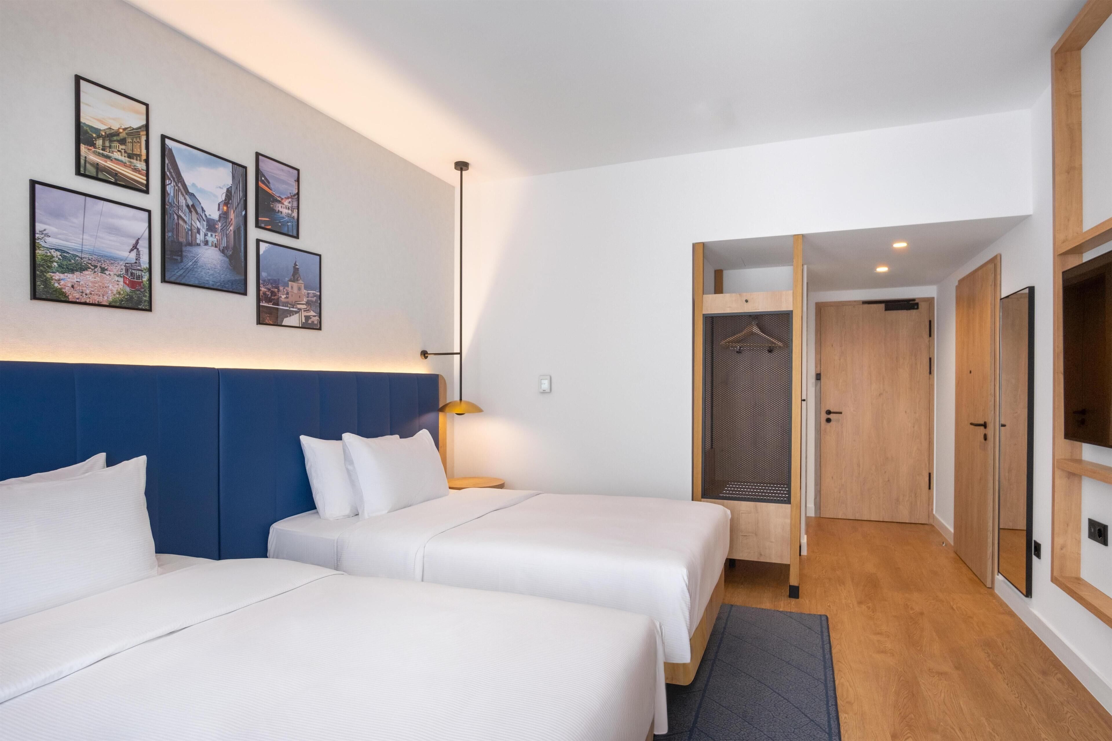 A hotel room with two beds, a blue headboard, a wall with framed pictures, a wooden wardrobe, and a hanging rack.