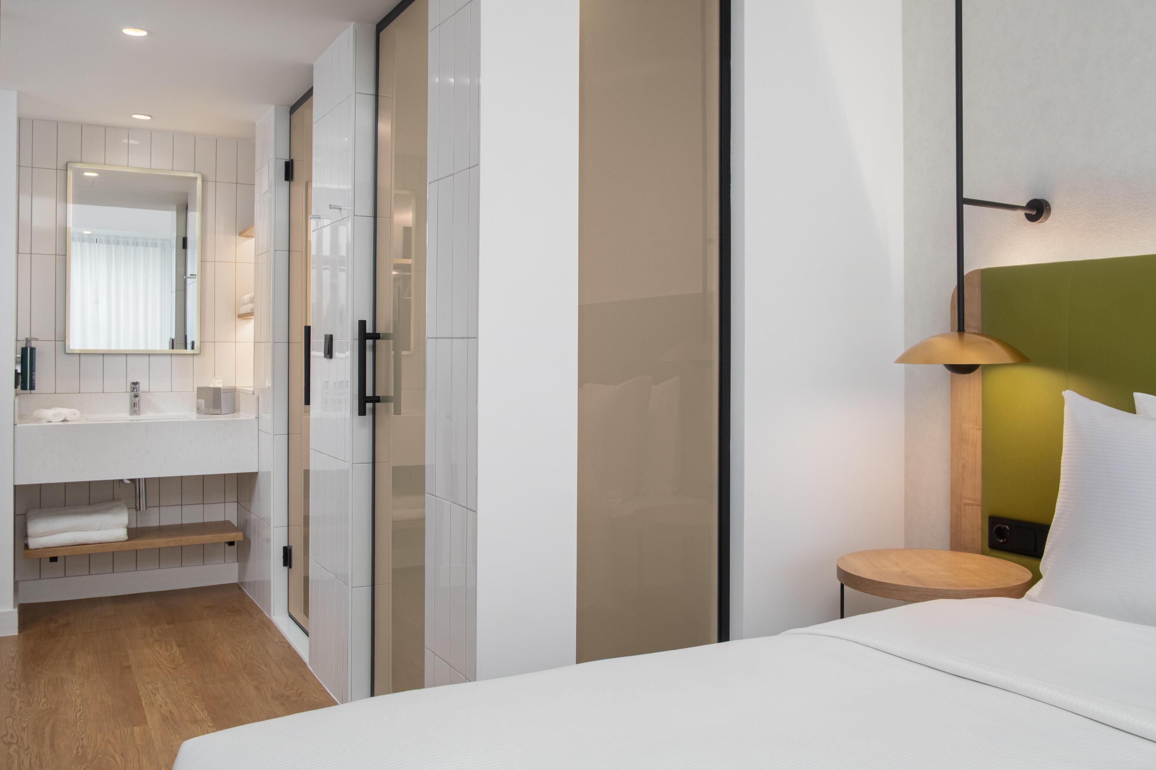A modern hotel room with a large mirror, a bedside table with a lamp, a wooden floor, and a built-in wardrobe.