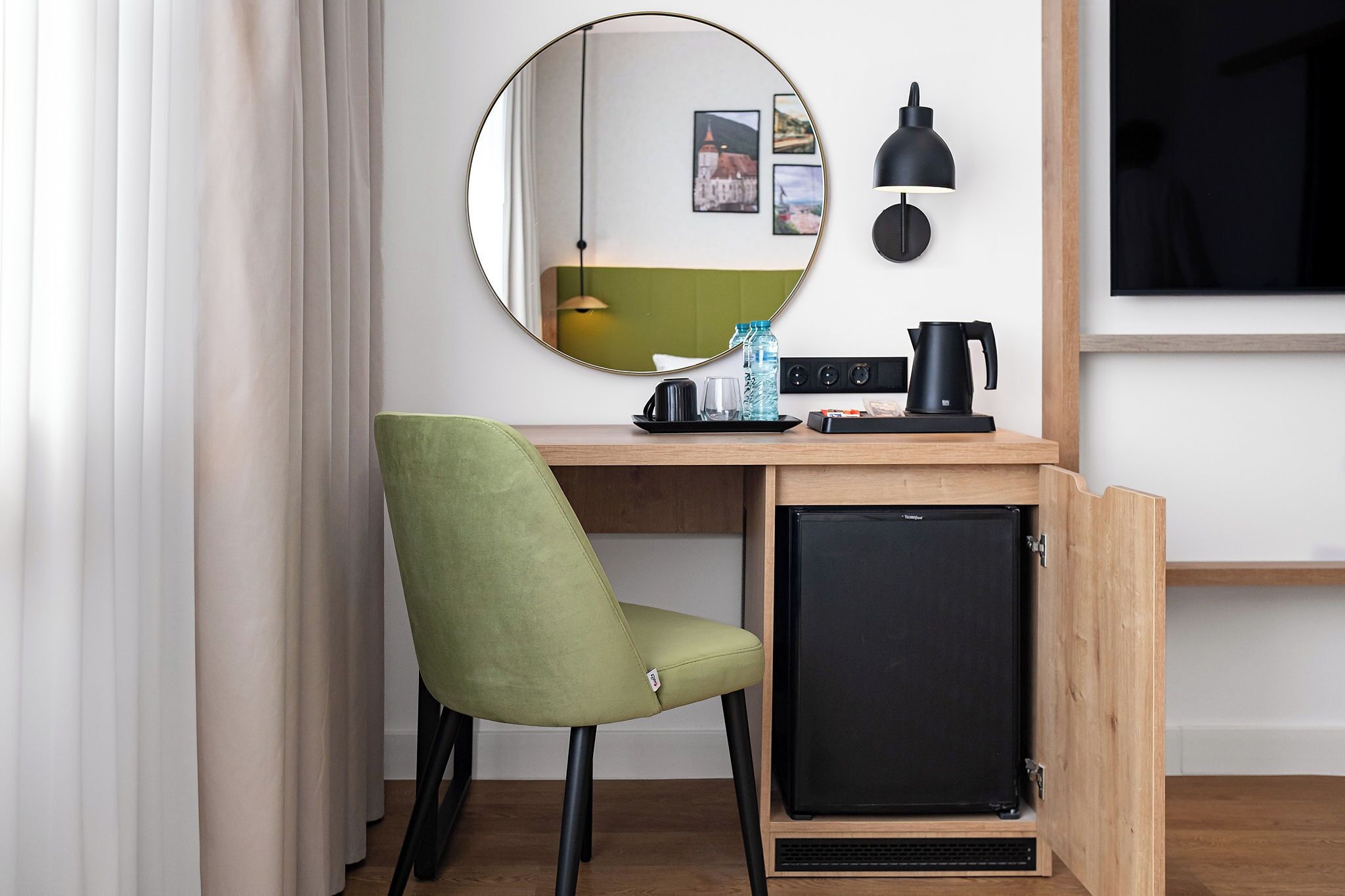 A compact room with a desk, a green chair, a small black refrigerator, a round mirror, a black lamp, and a wall-mounted TV.
