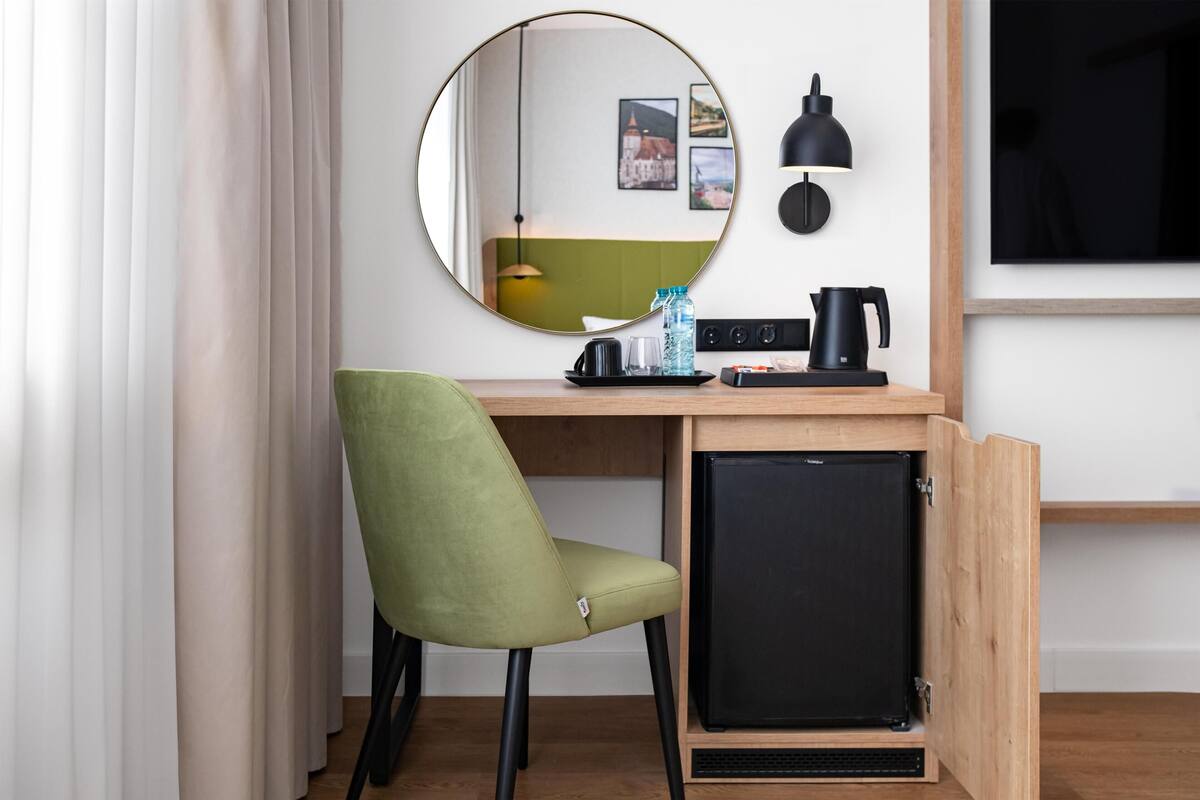 Une pièce de taille modeste, avec une chaise verte, un petit réfrigérateur noir, un miroir rond, une lampe noire et un téléviseur fixé au mur.