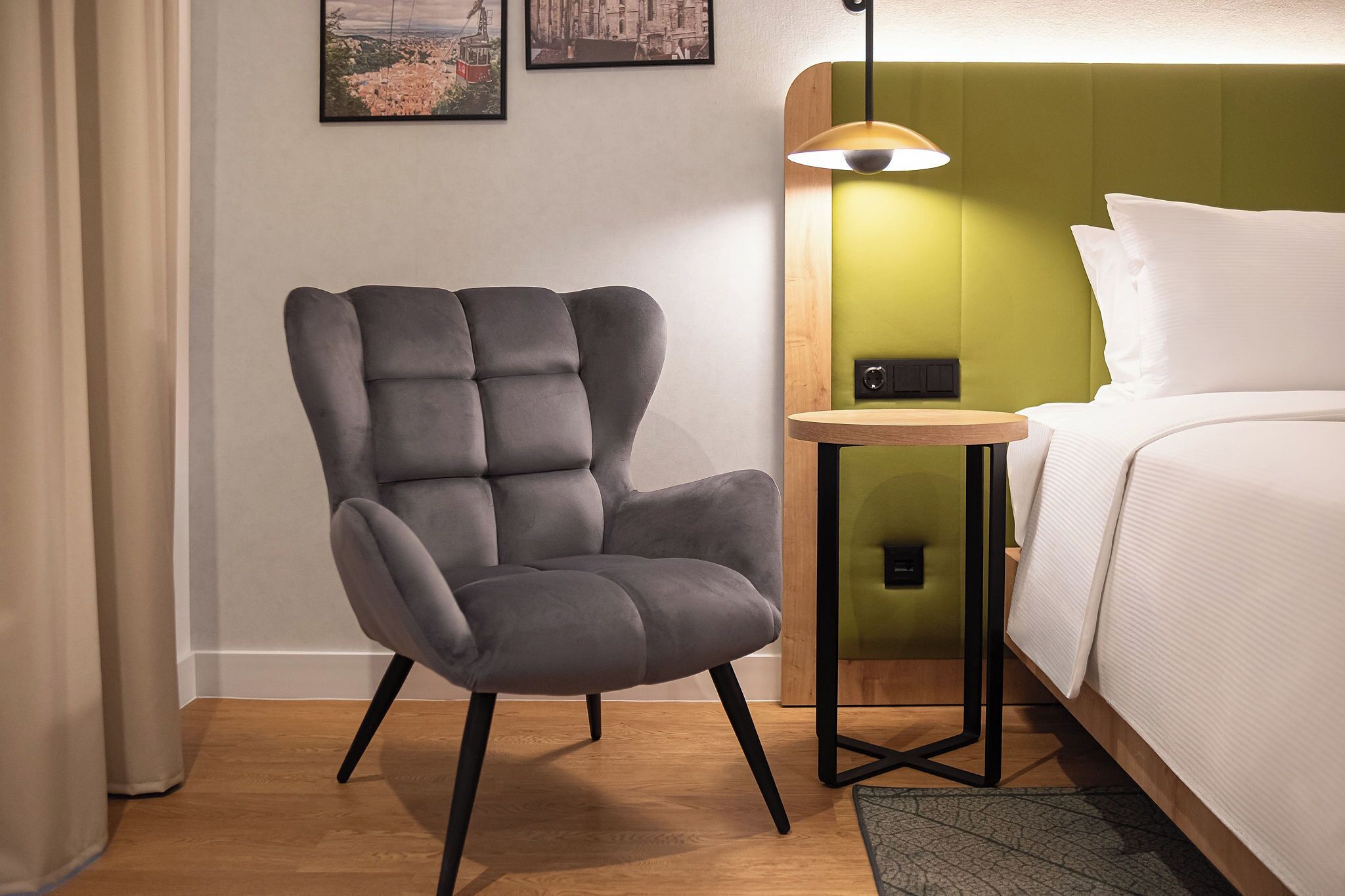 Una habitación de hotel moderna con un sillón tapizado gris, una mesita de madera, un cabecero verde y una cama con ropa blanca.