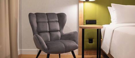 Una habitación de hotel moderna con un sillón tapizado gris, una mesita de madera, un cabecero verde y una cama con ropa blanca.