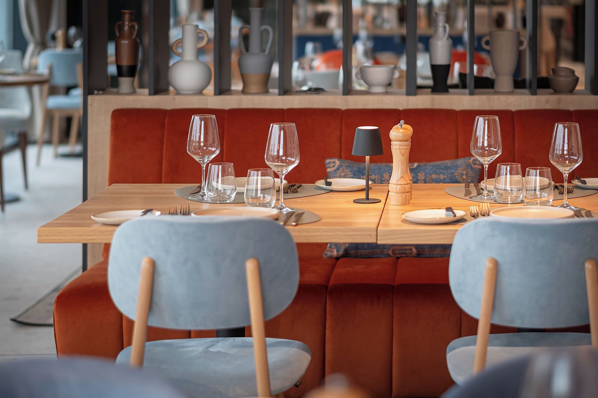 A restaurant setting with a wooden table set for two, two grey upholstered chairs, and a red cushioned bench.