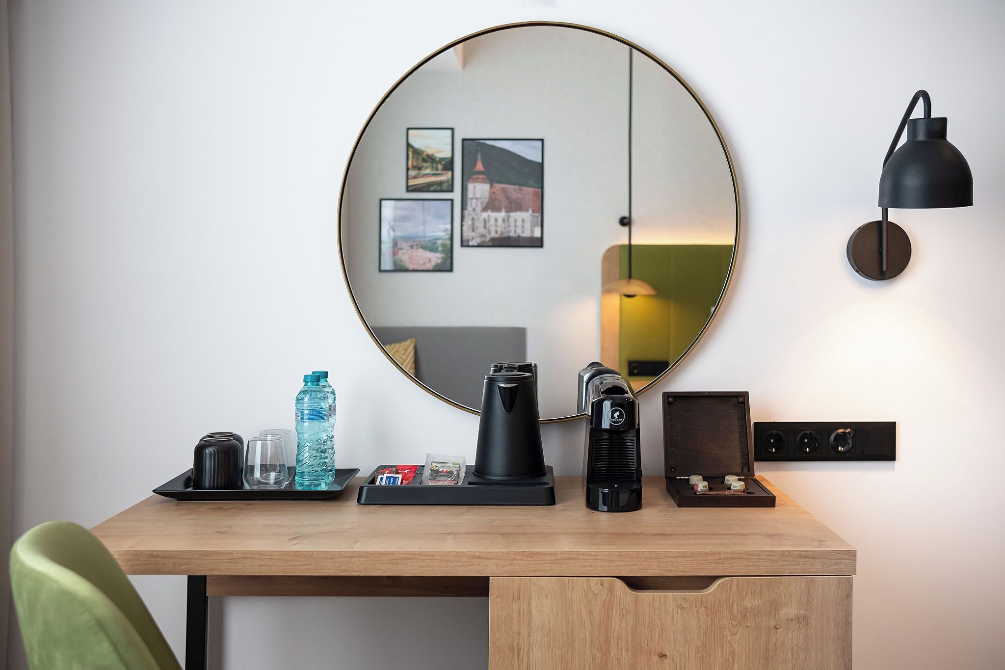 A wooden desk with a coffee maker, a kettle, a water bottle, and a tray with a glass and a cup. A round mirror hangs on the wall above the desk, reflecting a lamp and framed pictures.