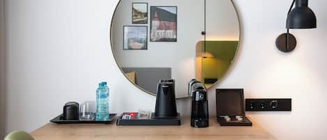 Escritorio de madera con una cafetera, un hervidor, una botella de agua y una bandeja con un vaso y una taza. Un espejo redondo cuelga en la pared sobre el escritorio, reflejando una lámpara y fotografías enmarcadas.