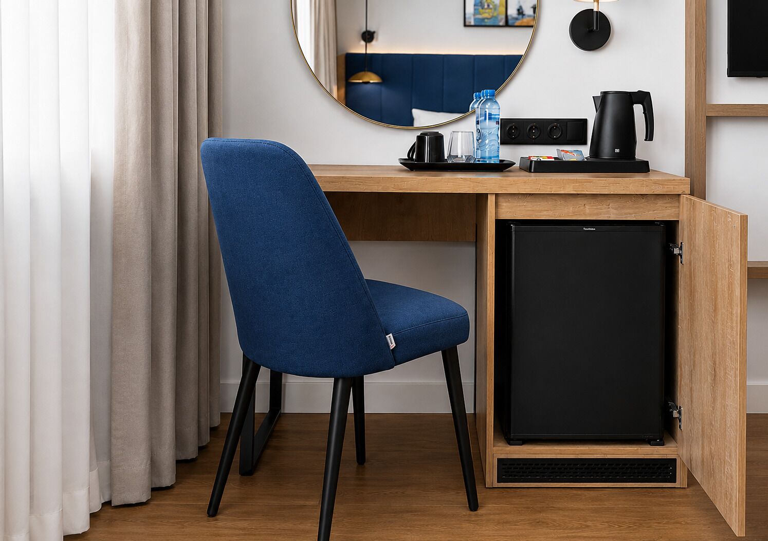 Habitación de hotel compacta con escritorio de madera, una silla azul, un refrigerador negro pequeño, una botella de agua, cafetera y televisión.