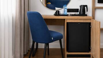 A compact hotel room with a wooden desk, a blue chair, a small black refrigerator, a water bottle, a coffee maker, and a television.