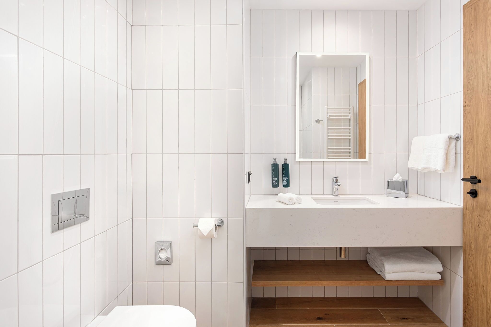 Un baño moderno con azulejos blancos, una puerta de madera, un lavamanos con espejo y un perchero para toallas.