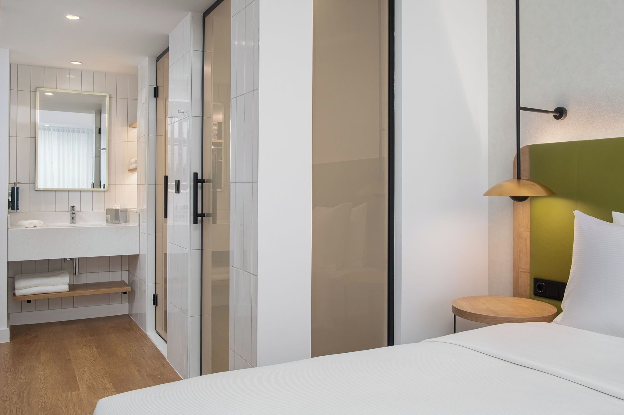 A modern hotel room with a large mirror, a bedside table with a lamp, a built-in wardrobe, and a wooden floor.