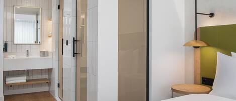 A modern hotel room with a large mirror, a bedside table with a lamp, a built-in wardrobe, and a wooden floor.