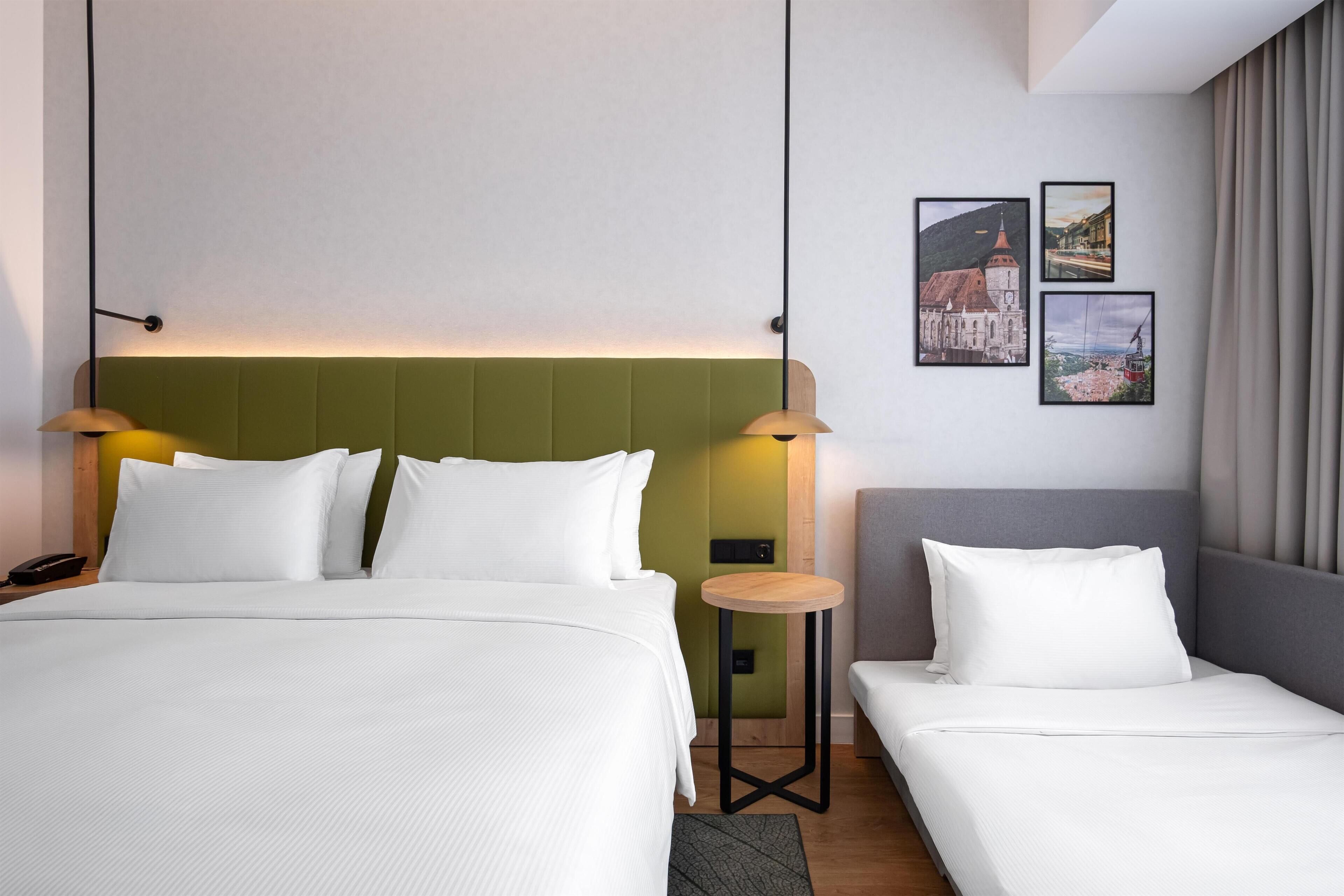 Habitación de hotel con una cama grande, una cama más pequeña, una mesita de noche, una lámpara y dos cuadros en la pared.