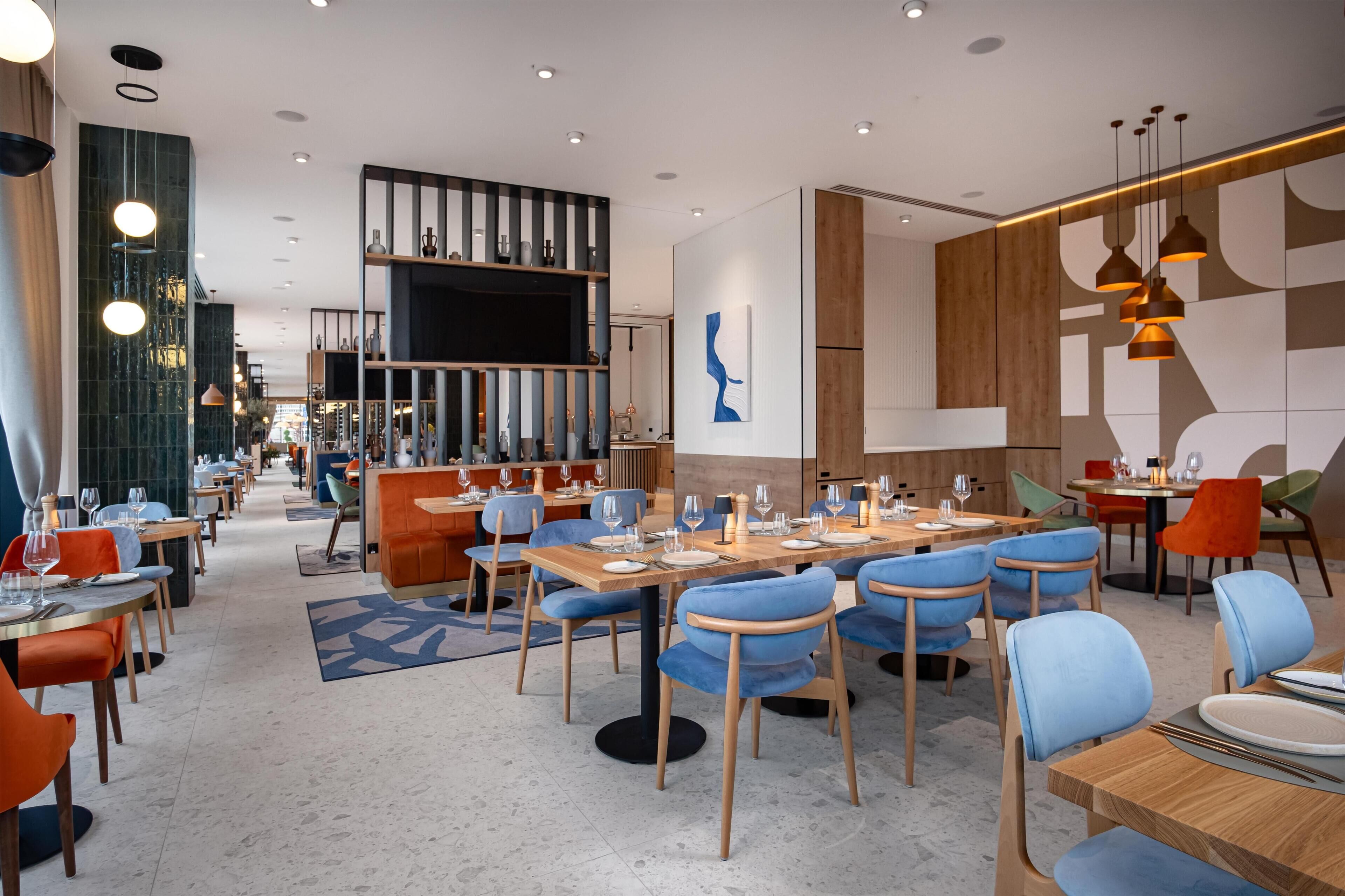 A modern restaurant interior with multiple dining tables, blue and orange chairs, and a bar area with shelves and a flat-screen TV.