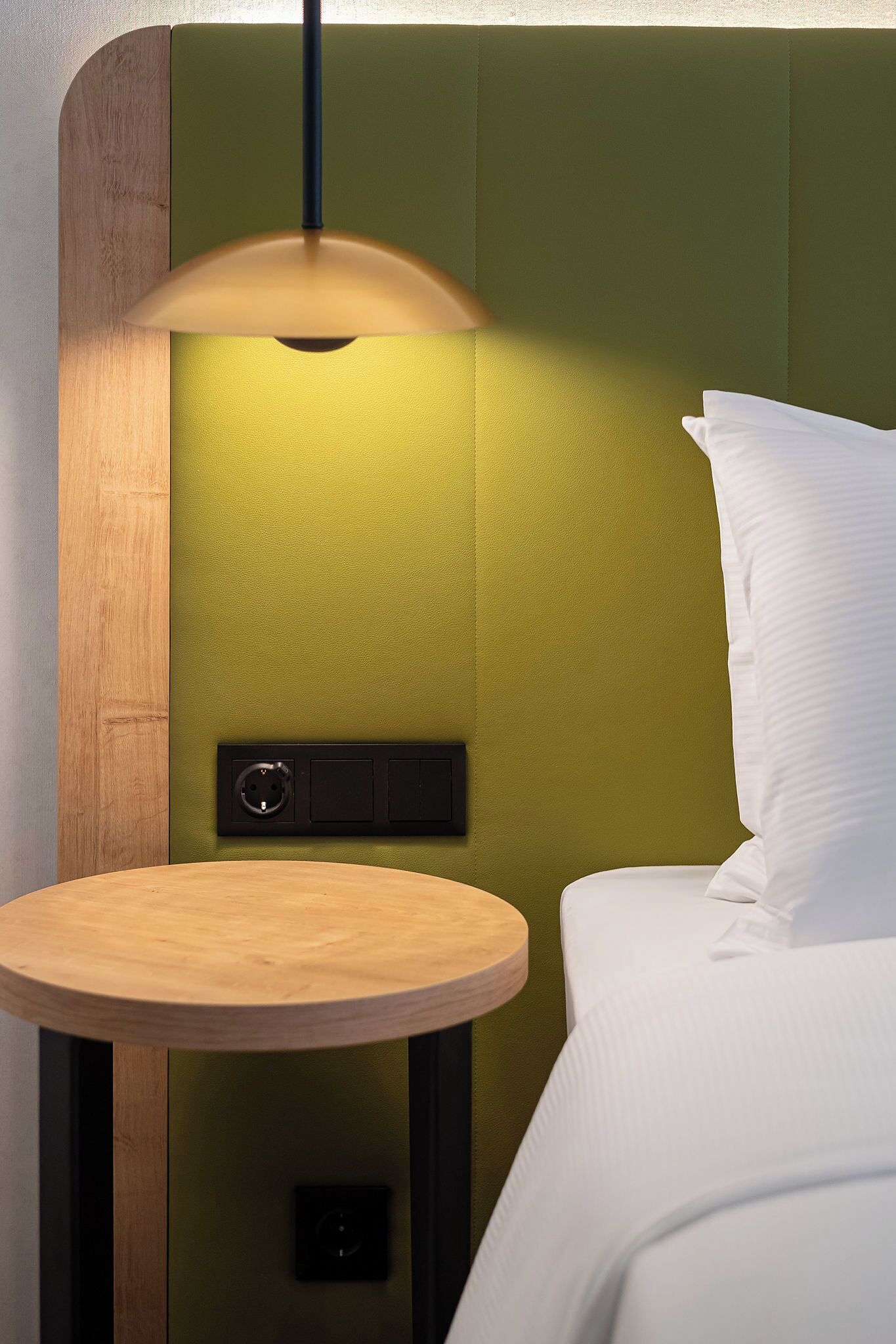 A bed with a white pillow, a round wooden table, and a wall-mounted light fixture.