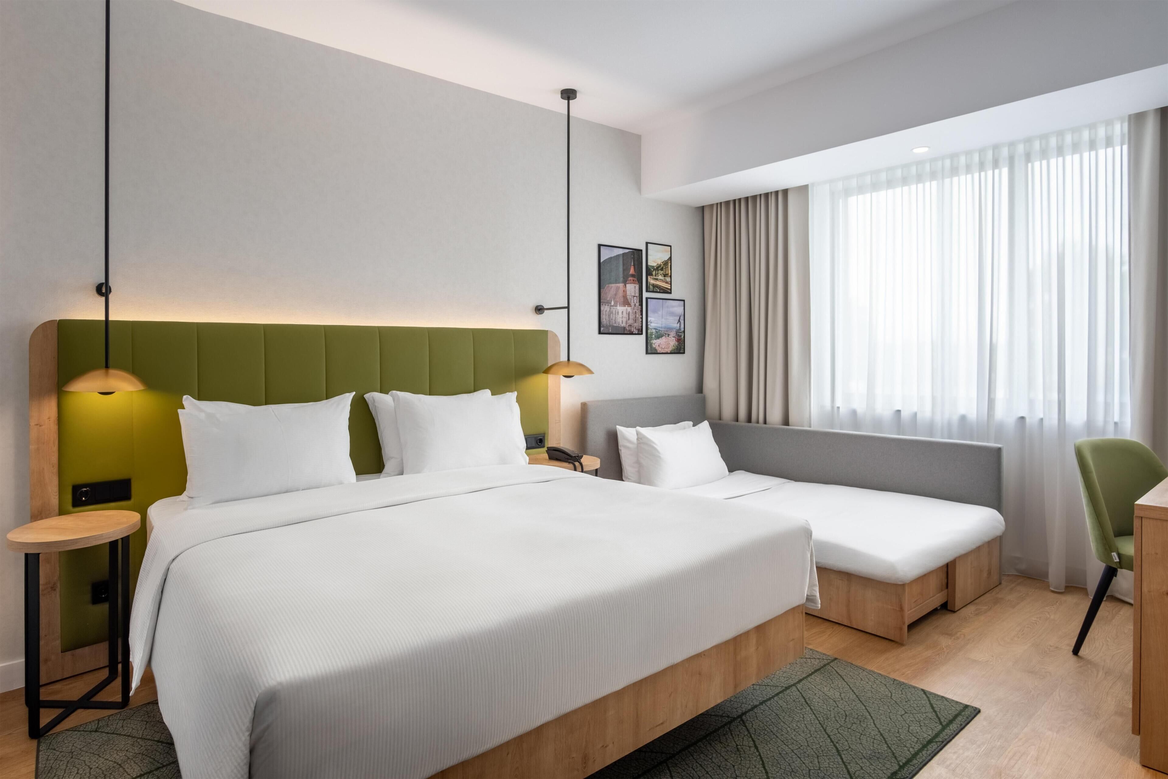 A hotel room with two beds, a green accent wall, a large window with curtains, and a small table.