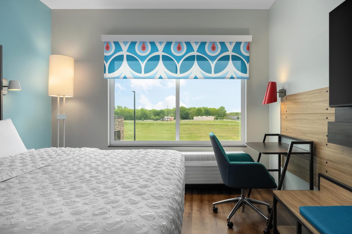 A hotel room with a large bed, a desk, a chair, and a window with a view of a green landscape.