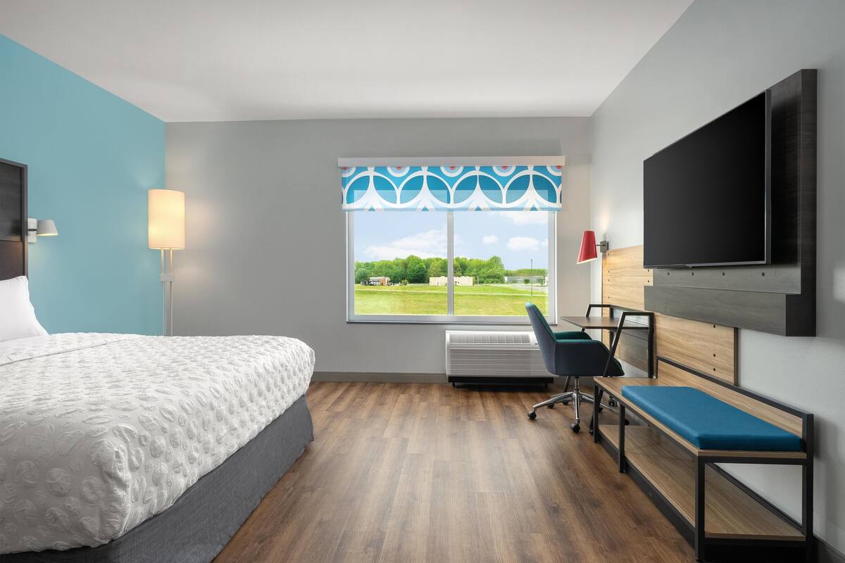 Habitación de hotel con cama, escritorio, silla, televisor y ventana con vista a follaje.