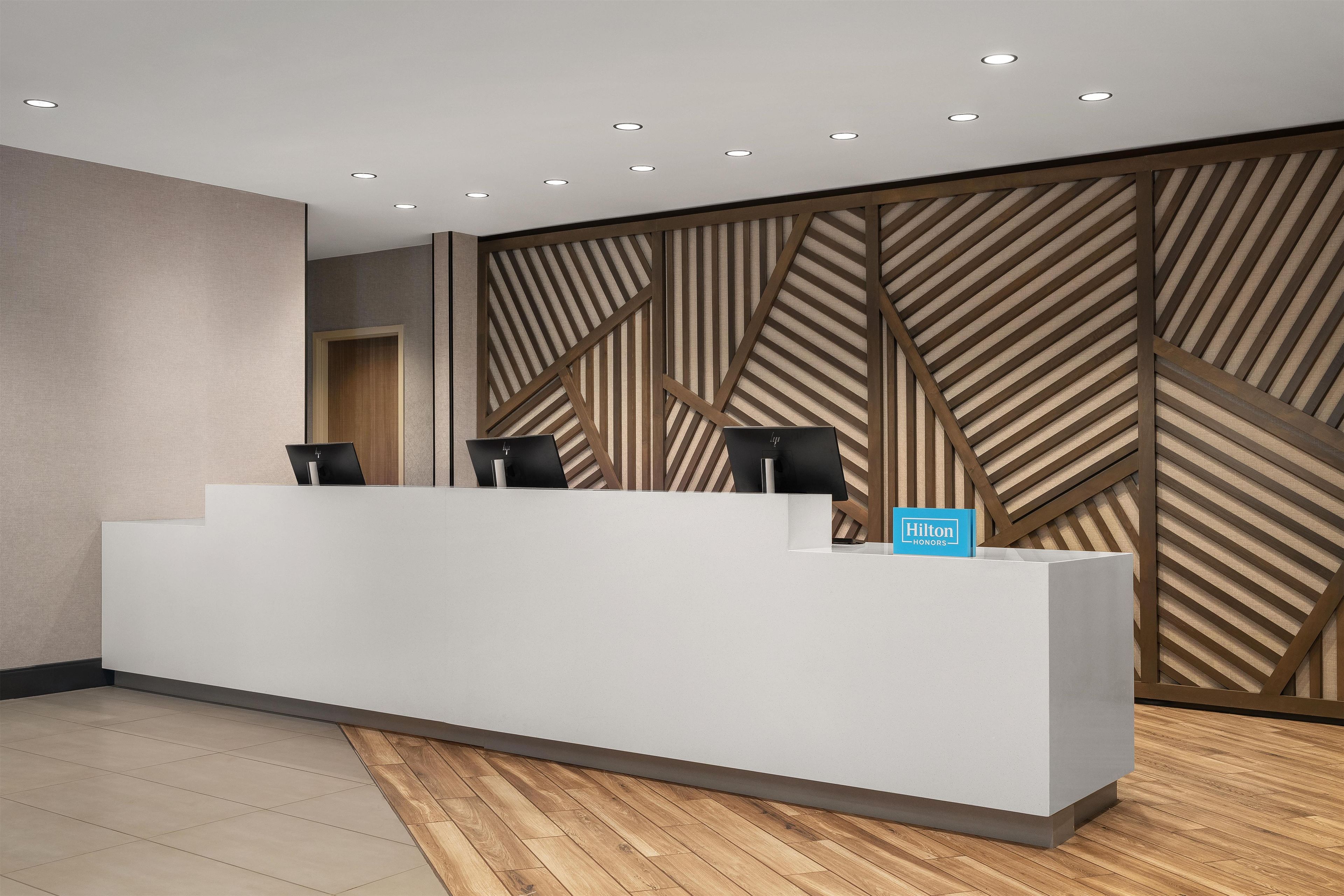A reception desk with a modern design, a sign indicating it's a Hilton Honors hotel, and a geometric patterned wall behind it.